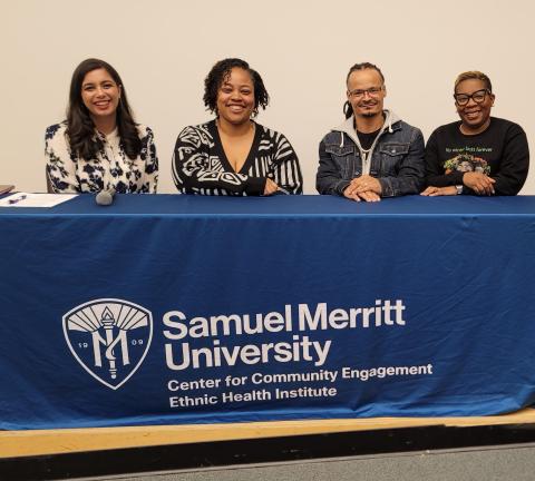Four individuals sit at a table with a blue Samuel Merritt University Center for Community Engagement, Ethnic Health Institute tablecloth.