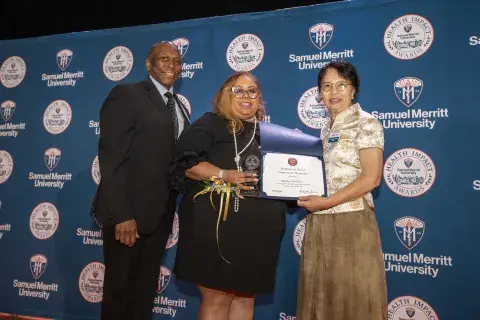 Photo of three individuals standing on stage at a ceremony. On the left is a man in a suit, in the middle is a woman holding an award and a certificate, and on the right is a woman in a traditional outfit holding a certificate. The background is a banner that says ‘Samuel Merritt University Health Impact Awards.'