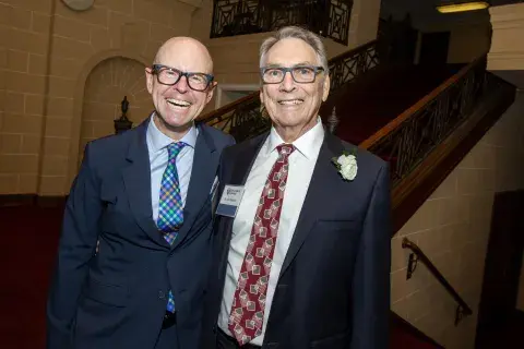 Photo of two men smiling and posing together. One is wearing a patterned tie and glasses, and the other is dressed in a black suit and red tie. They are standing in front of a staircase in a formal setting.