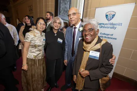 Photo of four individuals standing together. From left to right: a woman in a gold blouse and brown skirt, a woman in black, a man in a suit with a boutonnière, and a woman with white hair and glasses wearing a brown jacket. The background shows a banner for Samuel Merritt University.