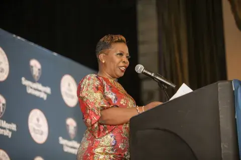 A woman in a dark dress with flowers standing at the podium while another man in a gray suit stands beside her. The background shows the ‘Samuel Merritt University Health Impact Awards’ logo.