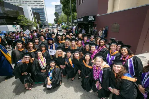 A large group of graduates in black caps and gowns gather together on a city street, smiling for a group photo. Many wear colorful leis and stoles, while faculty in academic regalia stand among them, holding program signs.