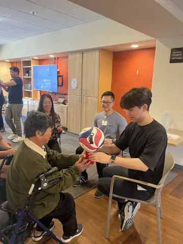 A young man in a black shirt sits facing an older woman using a walker, helping her hold a red, white, and blue basketball in a therapy exercise. Other students and participants are visible in the background, smiling and engaging in activities.