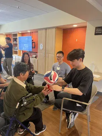 A young man in a black shirt sits facing an older woman using a walker, helping her hold a red, white, and blue basketball in a therapy exercise. Other students and participants are visible in the background, smiling and engaging in activities.