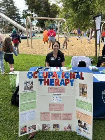 A woman wearing a black Samuel Merritt University polo shirt sits at a table in a park with a colorful trifold board titled “Occupational Therapy.” The board features information about the profession, application requirements, and activities of daily living. A playground with children and families is visible in the background.
