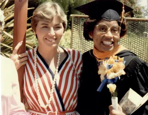 Two women stand close together outdoors, smiling at the camera. One wears a red-and-white striped dress with a beaded necklace, while the other is dressed in a black graduation cap and gown, holding flowers and a diploma.