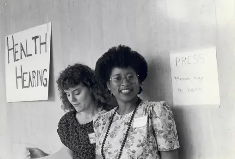 Black-and-white photo of two women standing by handmade signs that read “HEALTH HEARING” and “PRESS Please sign in here.” One woman looks down at papers, while the other smiles at the camera, wearing glasses and a floral dress.