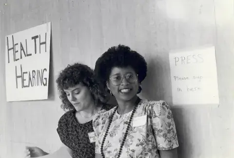 Black-and-white photo of two women standing by handmade signs that read “HEALTH HEARING” and “PRESS Please sign in here.” One woman looks down at papers, while the other smiles at the camera, wearing glasses and a floral dress.