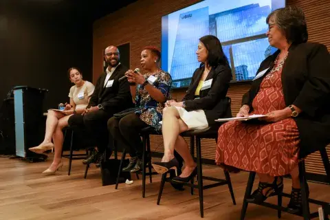 Five people sitting on a panel, one holding microphone and speaking.