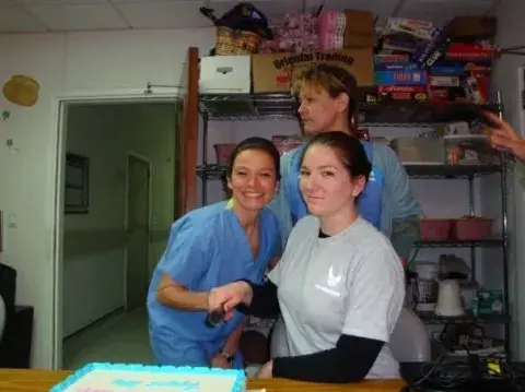 Three healthcare professionals smile around a table with a decorated cake in a break room filled with board games and supplies.