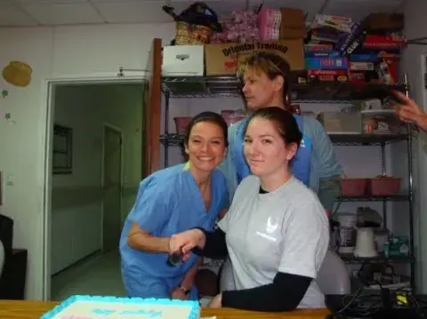 Three healthcare professionals smile around a table with a decorated cake in a break room filled with board games and supplies.