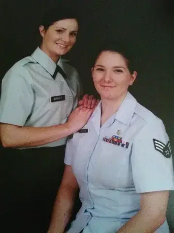 Two uniformed Air Force service members pose for a formal studio portrait, one standing with a hand on the other’s shoulder.