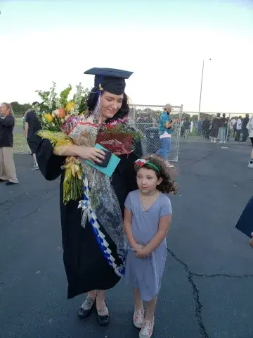 A graduate wearing a cap and gown holds several bouquets while standing beside a young girl in a gray dress at an outdoor ceremony.
