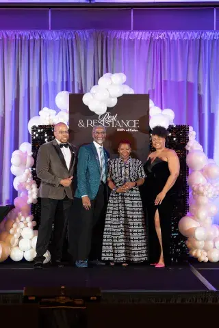 Four people stand on stage in formal attire in front of a backdrop that reads “Rest & Resistance: DNPs of Color.” The backdrop is decorated with white balloons and purple lighting.
