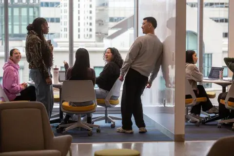Five students sitting around a table, talking and laughing, in front of a large window 