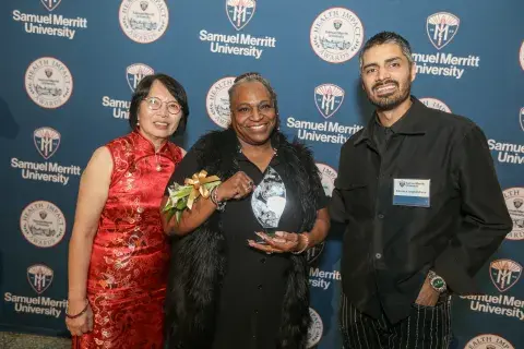 Three individuals pose in front of a Samuel Merritt University Health Impact Awards backdrop, with one person holding an award.