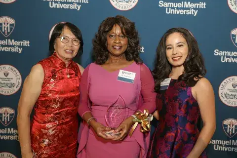 Three individuals pose in front of a Samuel Merritt University Health Impact Awards backdrop, with the person in the center holding an award.