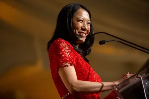 A woman in a red lace dress smiles while speaking at a podium with a microphone during a formal event.