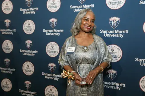A woman poses in front of a Samuel Merritt University Health Impact Awards backdrop, holding an award.