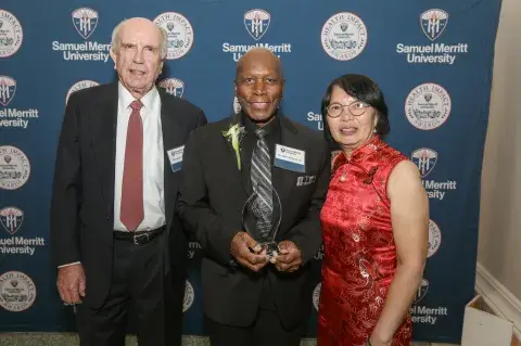 Three individuals pose at the Samuel Merritt University Health Impact Awards, with the person in the center holding an award.