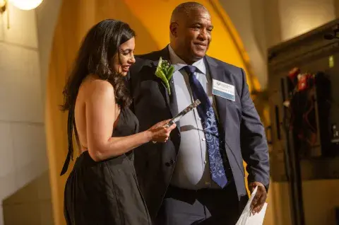 Two individuals stand together on stage at the Samuel Merritt University Health Impact Awards, with one holding an award.