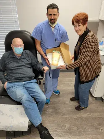 Three people smile while holding a cake shaped like a foot inside a medical office.