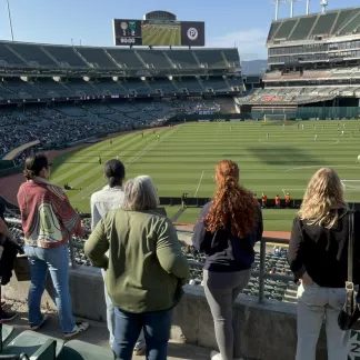 Six women stand at the railing of the Oakland Coliseum during a Soul SC match.