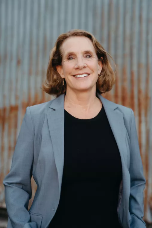 Dean of Nursing smiling, wearing a black top and grey blazer against a rusted metal background.
