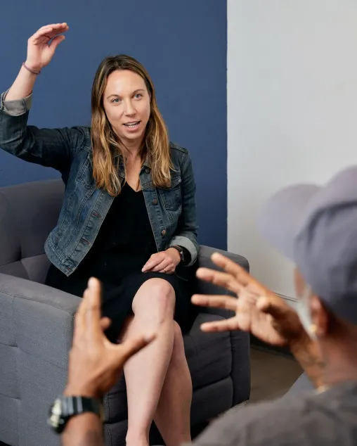 Student wearing a black dress and denim jacket holder their hand up over their head against a blue background. Another person in the foreground gestures with both their hands.