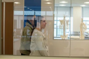 Two students walk through hallway with skeleton in classroom behind window