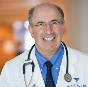 Portrait of a smiling male physician wearing glasses, a white lab coat, blue shirt, and tie with a stethoscope around his neck.