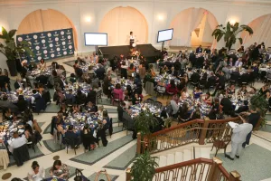 A bird’s-eye view of a formal awards event shows attendees seated at round tables during dinner as a speaker addresses the room from a stage at the front.