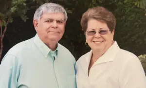 Portrait of a smiling couple standing outdoors in front of greenery, dressed in light-colored clothing.