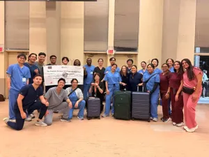 Group photo of students in scrubs with two suitcases in front of them and a banner behind