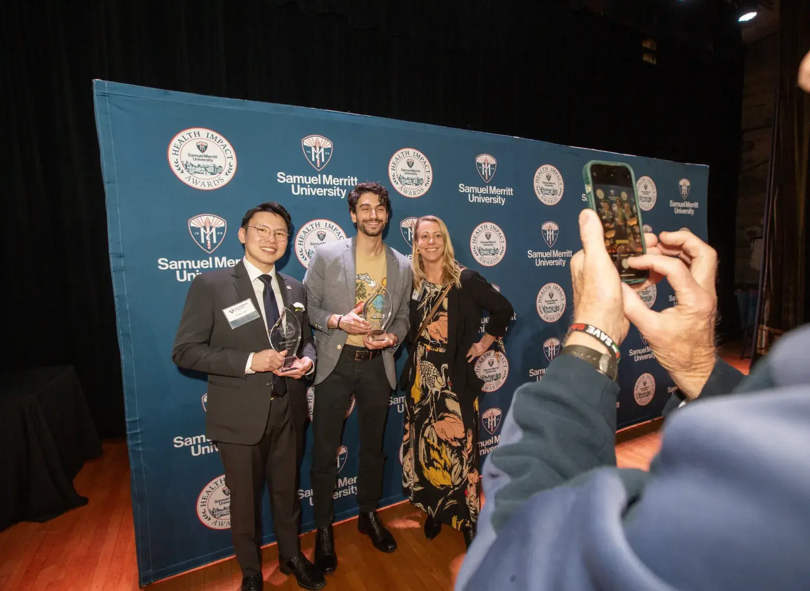 Three Health Impact Award attendees pose in front of a Samuel Merritt University step-and-repeat backdrop, holding glass awards, while someone in the foreground takes their photo with a smartphone.