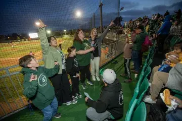 A lively group of Samuel Merritt University attendees, including children and adults in Oakland Ballers gear, cheer from the stands during the game.