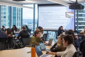 Students work in small groups with laptops during a class session at Samuel Merritt University, with a presentation on legal and ethical issues in healthcare projected at the front of the room.