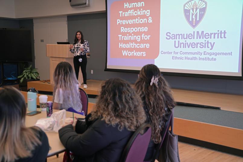 Sam Alavi-Irvine stands at podium in front of audience. Screen behind her reads: Human Trafficking Prevention & Response Training for Healthcare Workers. Samuel Merritt University Center for Community Engagement, Ethnic Health Institute