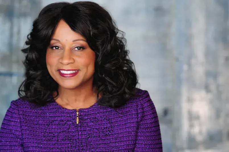 Paulina Van with styled black hair and a bright smile poses against a soft blue-gray background, wearing a textured purple jacket with a gold zipper detai