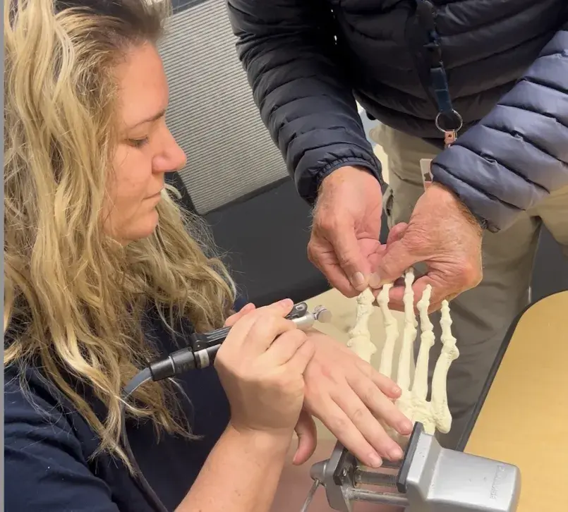 A student practices podiatric techniques using a skeletal model of a foot held steady in a vise, guided by an instructor standing beside her.