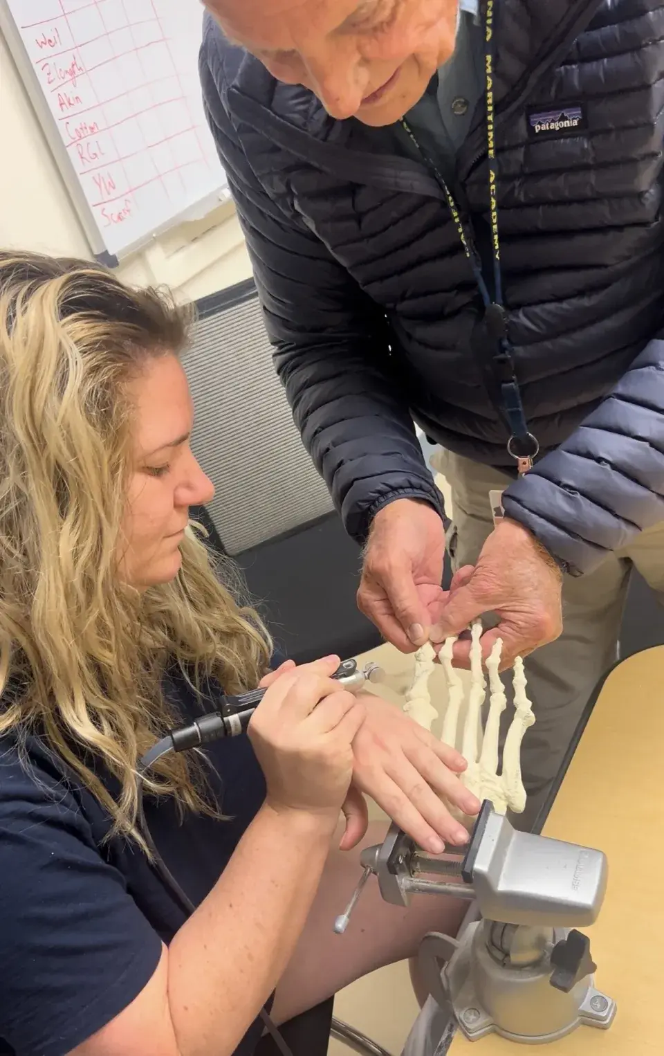 A student practices podiatric techniques using a skeletal model of a foot held steady in a vise, guided by an instructor standing beside her.