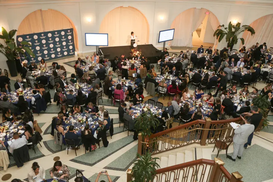 A bird’s-eye view of a formal awards event shows attendees seated at round tables during dinner as a speaker addresses the room from a stage at the front.
